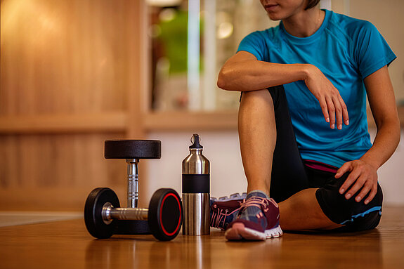 Eine Frau sitzt auf dem Boden eines Fitnessraums mit Hanteln und Trinkflasche.
