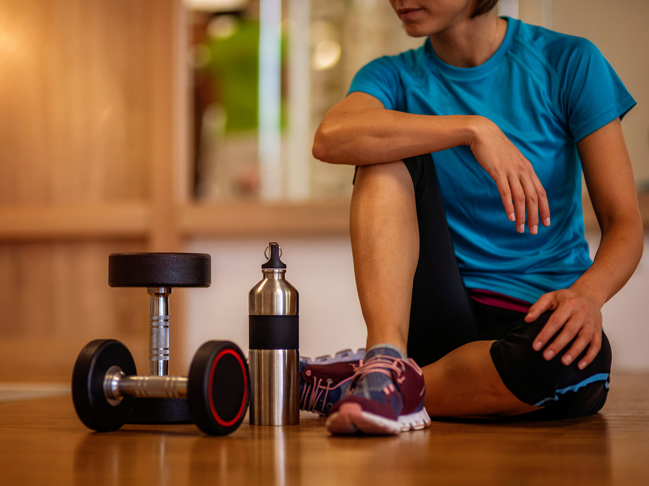 Eine Frau sitzt auf dem Boden eines Fitnessraums mit Hanteln und Trinkflasche.