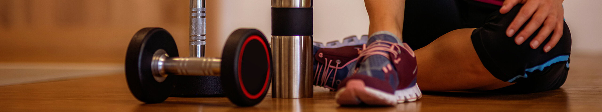 Eine Frau sitzt auf dem Boden eines Fitnessraums mit Hanteln und Trinkflasche.