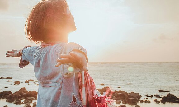 Eine Frau atmet tief ein mit ausgebreiteten Händen am Meer.
