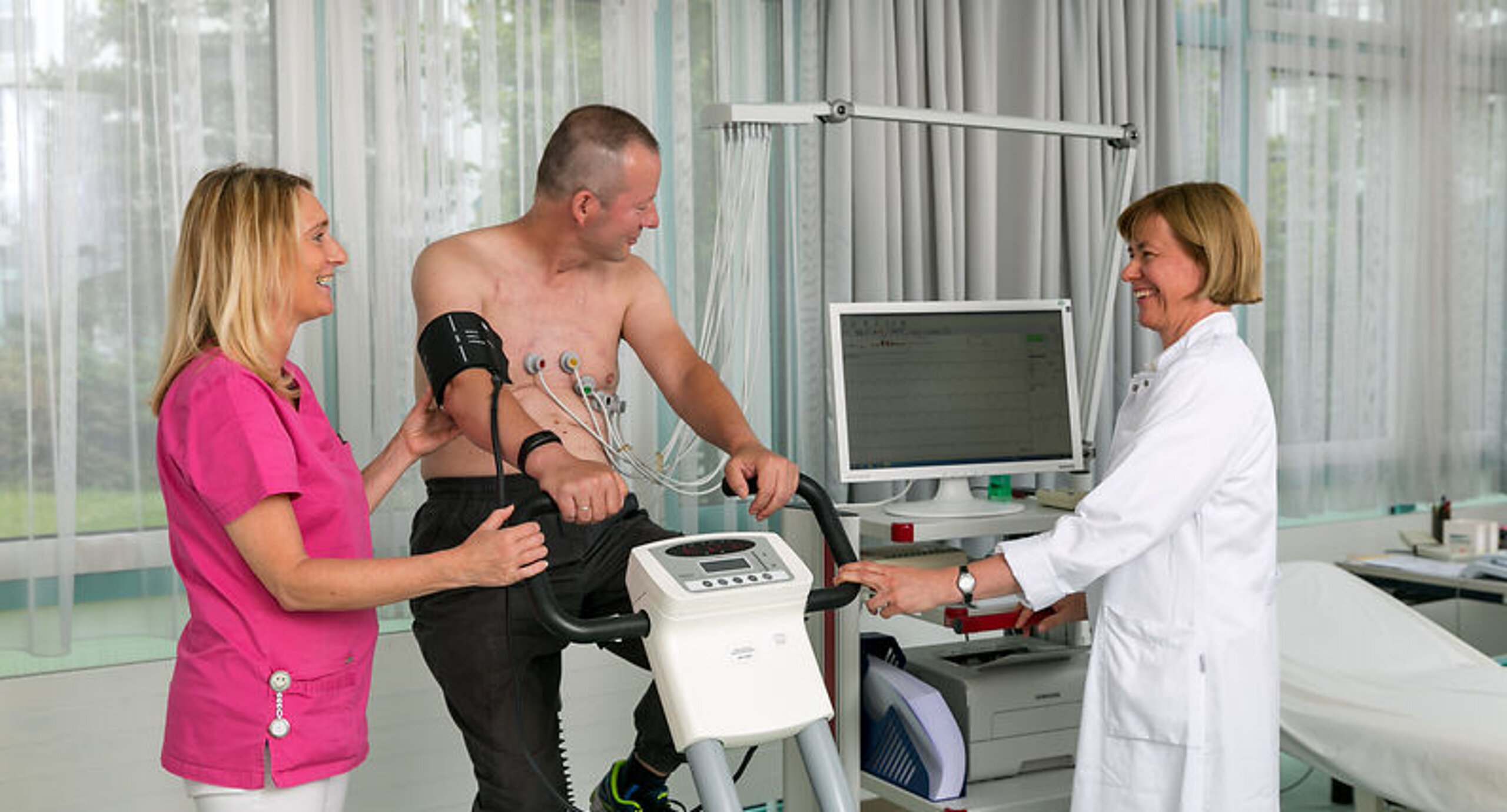 Belastungs-EKG Ein Patient sitzt auf einem Fahrradergometer, links neben ihm steht eine Frau in rosafarbenem Kittel, rechts neben ihm eine Ärztin in weißem Kittel am PC.