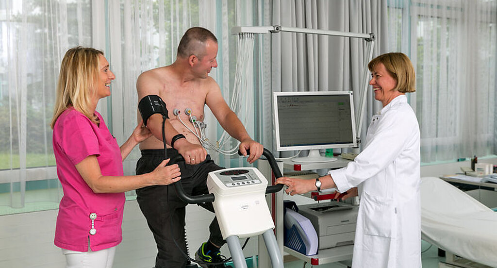 Belastungs-EKG Ein Patient sitzt auf einem Fahrradergometer, links neben ihm steht eine Frau in rosafarbenem Kittel, rechts neben ihm eine Ärztin in weißem Kittel am PC.