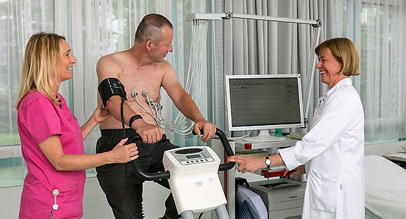 Ein Patient sitzt auf einem Fahrradergometer, links neben ihm steht eine Frau in rosafarbenem Kittel, rechts neben ihm eine Ärztin in weißem Kittel am PC.