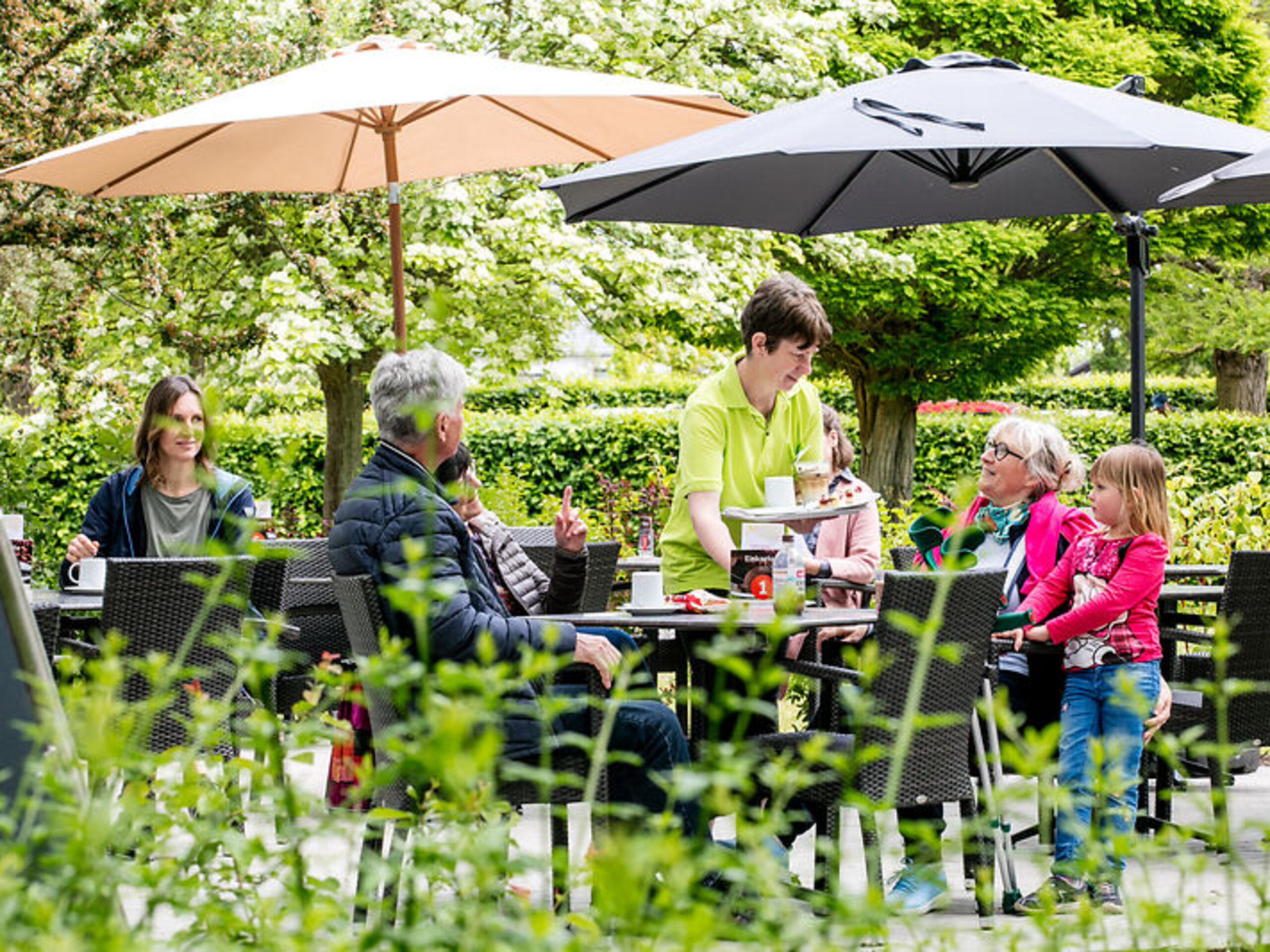 Auf einer Terrasse im Grünen sitzen Menschen an Tischen, eine Familie wird von einer Servicekraft bedient.