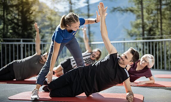 Eine Trainerin korrigiert einen Mann der in einer seitlichen Yogapose ist auf einer roten Matte