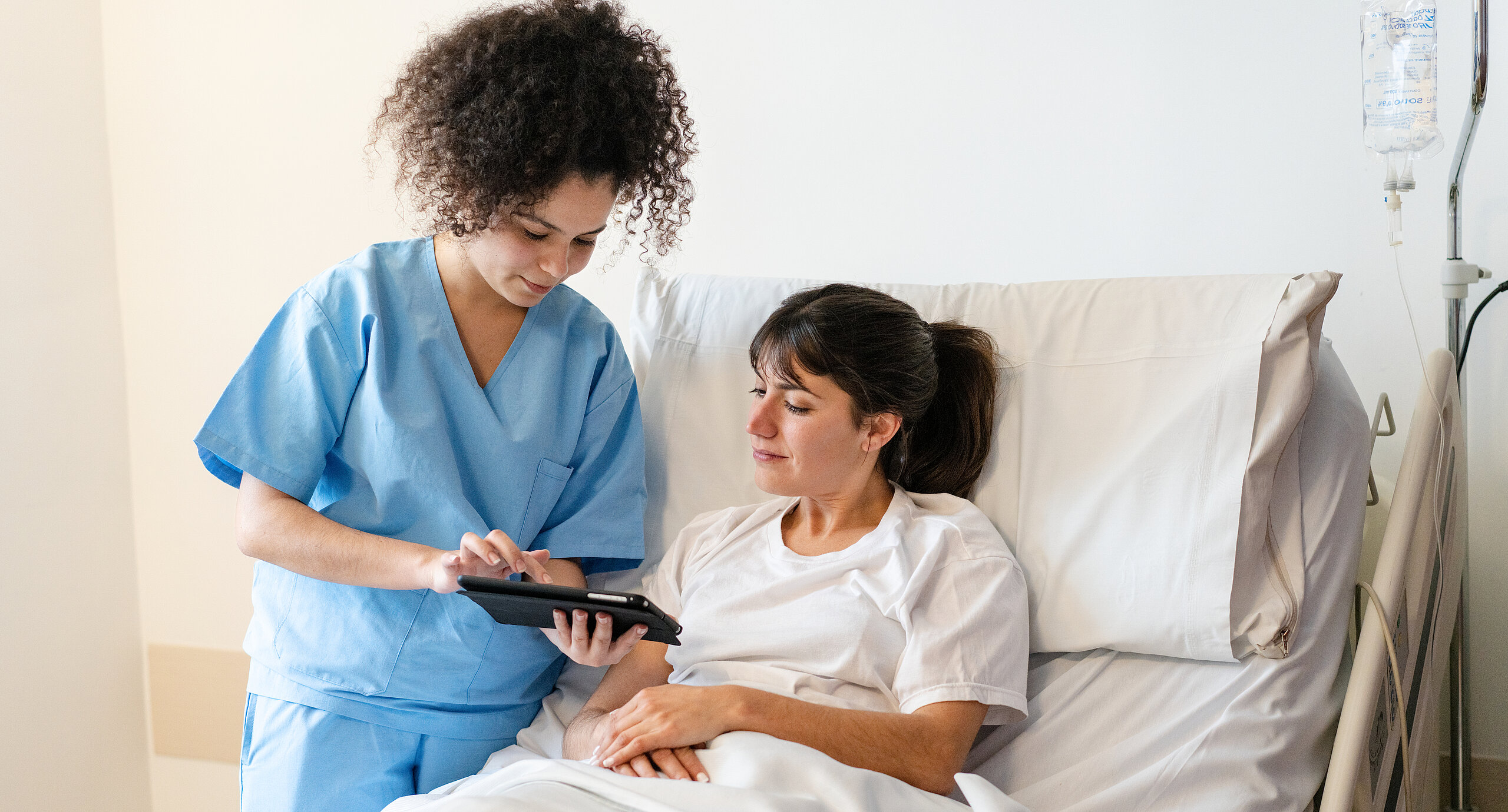 Eine Pflegekraft in blauer Kleidung zeigt einer Patientin im Krankenhausbett etwas auf einem Tablet.