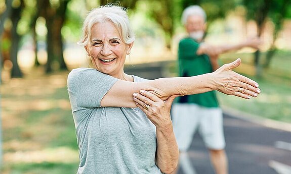 Frau macht Arm-Dehnübungen im Park und lächelt dabei freundlich.