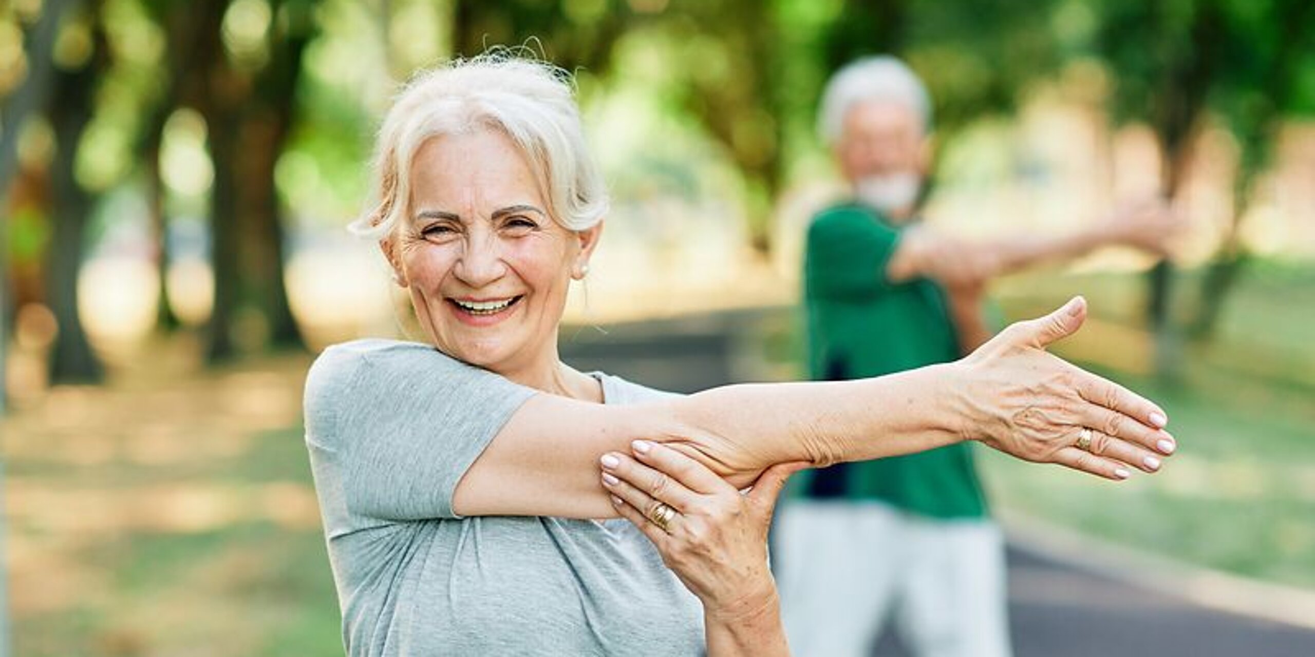 Frau macht Arm-Dehnübungen im Park und lächelt dabei freundlich.