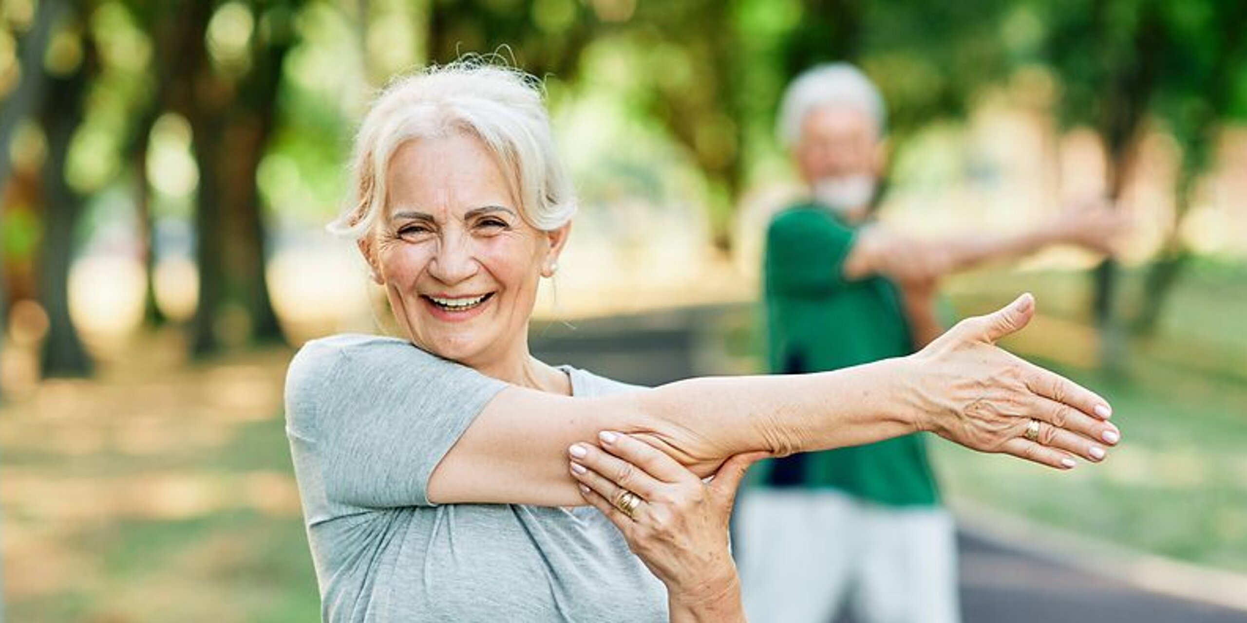 Frau macht Arm-Dehnübungen im Park und lächelt dabei freundlich.