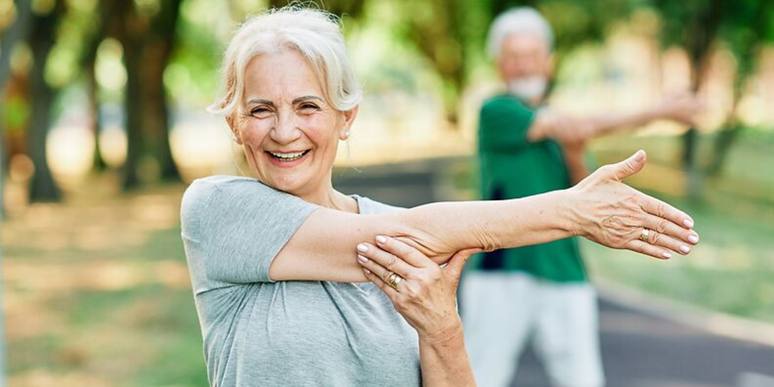 Frau macht Arm-Dehnübungen im Park und lächelt dabei freundlich.