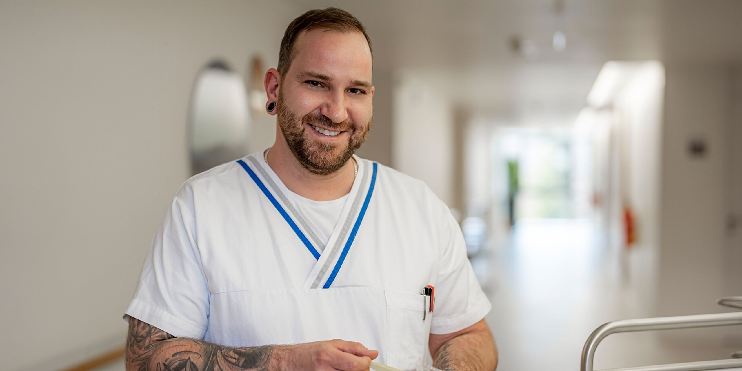 Ein lächelnder Pfleger mit Tattoos steht im Gang einer Klinik.