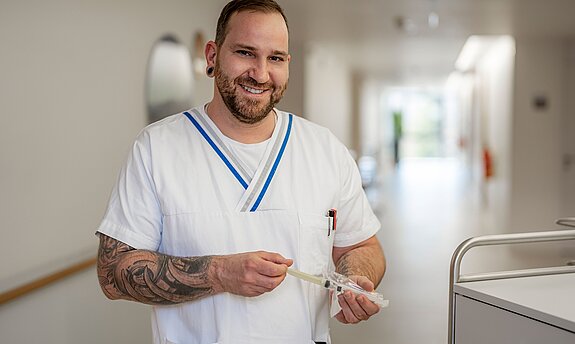 Ein lächelnder Pfleger mit Tattoos steht im Gang einer Klinik.