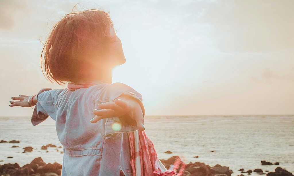 Eine Frau atmet tief ein mit ausgebreiteten Händen am Meer.
