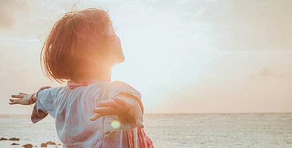 Eine Frau atmet tief ein mit ausgebreiteten Händen am Meer.