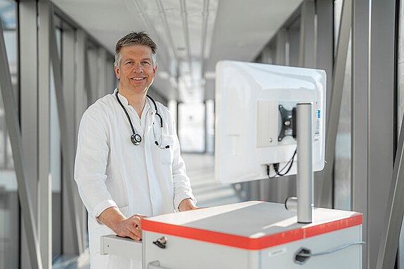 Ein lächelnder Arzt mit Visitenwagen im Gang.