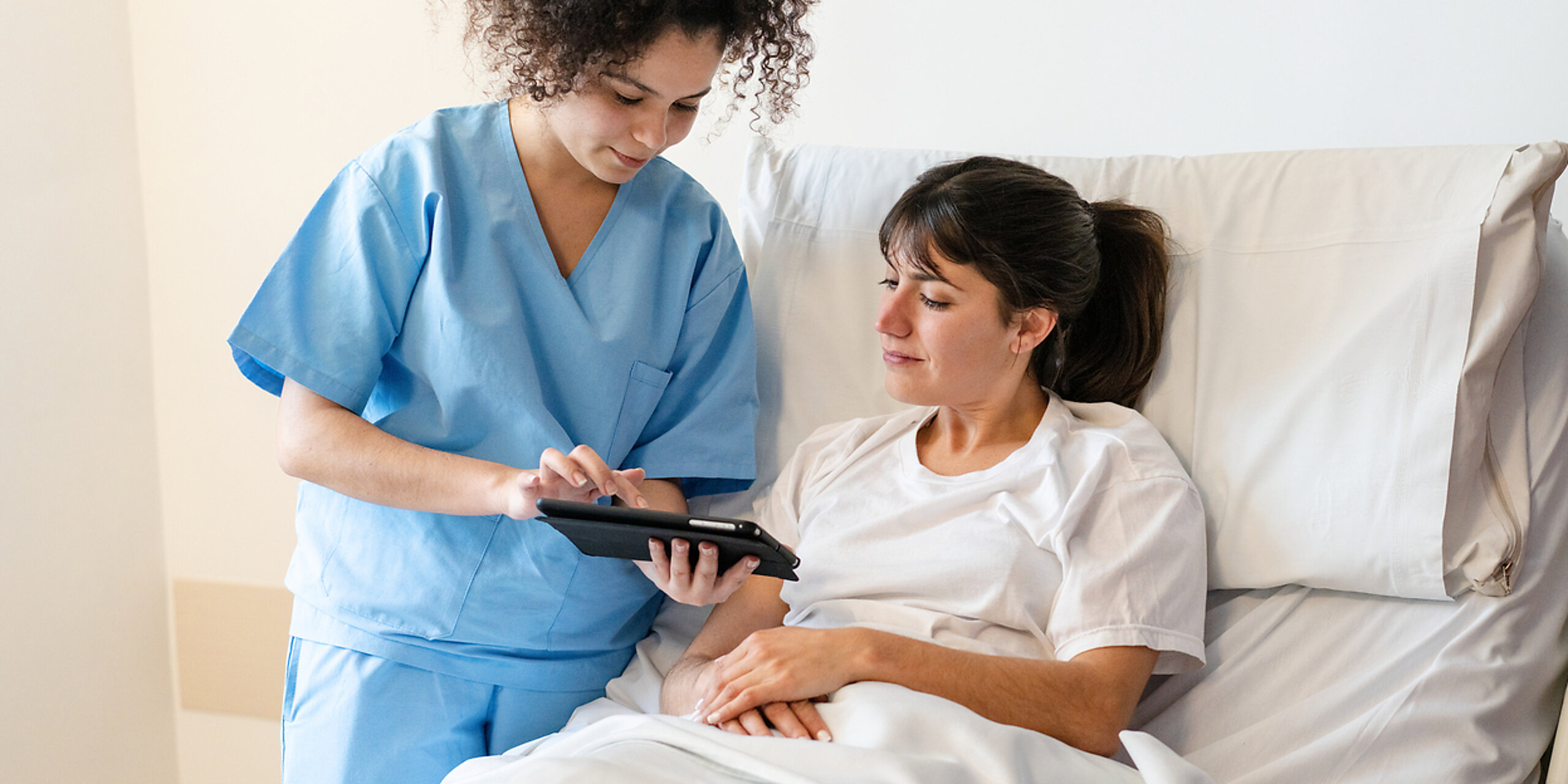 Eine Pflegerin zeigt einer Patientin im Krankenhausbett etwas auf dem Tablet.
