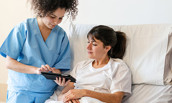 Eine Pflegerin zeigt einer Patientin im Krankenhausbett etwas auf dem Tablet.