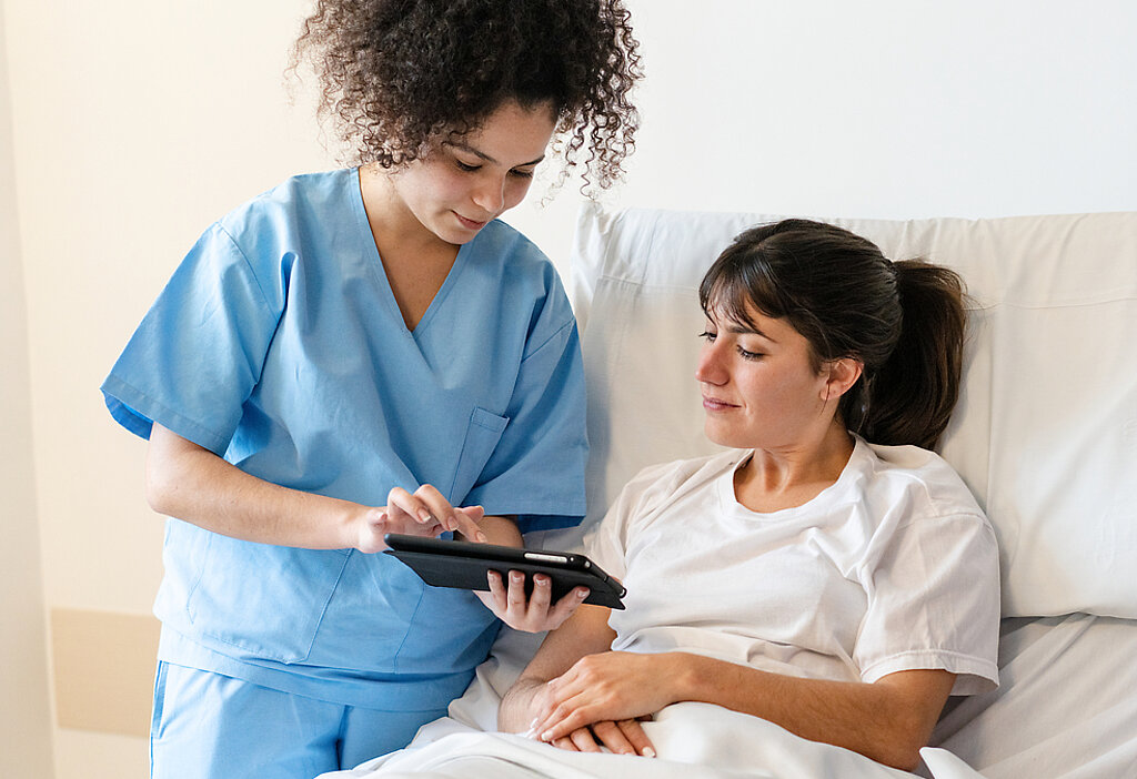 Eine Pflegerin zeigt einer Patientin im Krankenhausbett etwas auf dem Tablet.