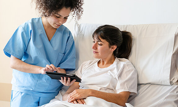 Eine Pflegerin zeigt einer Patientin im Krankenhausbett etwas auf dem Tablet.