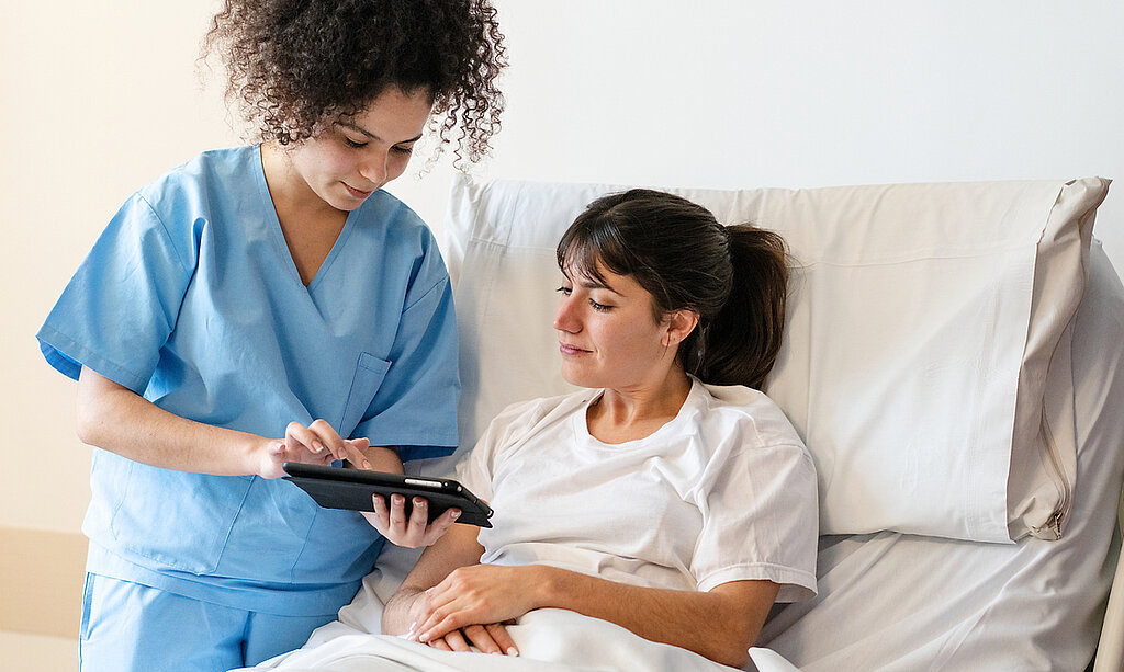 Eine Pflegerin zeigt einer Patientin im Krankenhausbett etwas auf dem Tablet.
