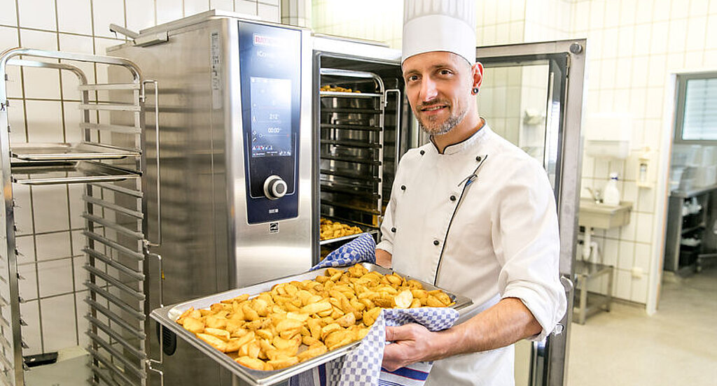 Ein Koch steht vor einem Industriebackofen und lächelt mit einem Blech Kartoffeln in den Händen in die Kamera.