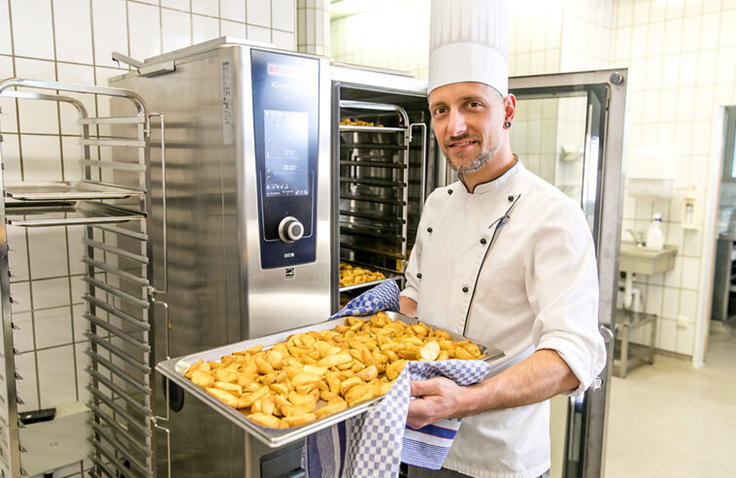 Küche Ein Koch steht vor einem Industriebackofen und lächelt mit einem Blech Kartoffeln in den Händen in die Kamera.