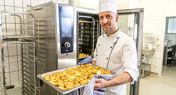 Ein Koch steht vor einem Industriebackofen und lächelt mit einem Blech Kartoffeln in den Händen in die Kamera.
