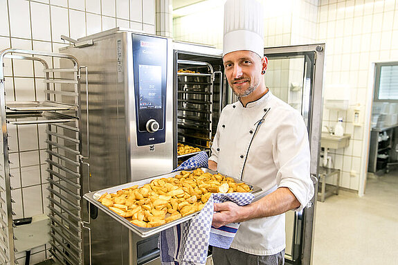 Küche Ein Koch steht vor einem Industriebackofen und lächelt mit einem Blech Kartoffeln in den Händen in die Kamera.
