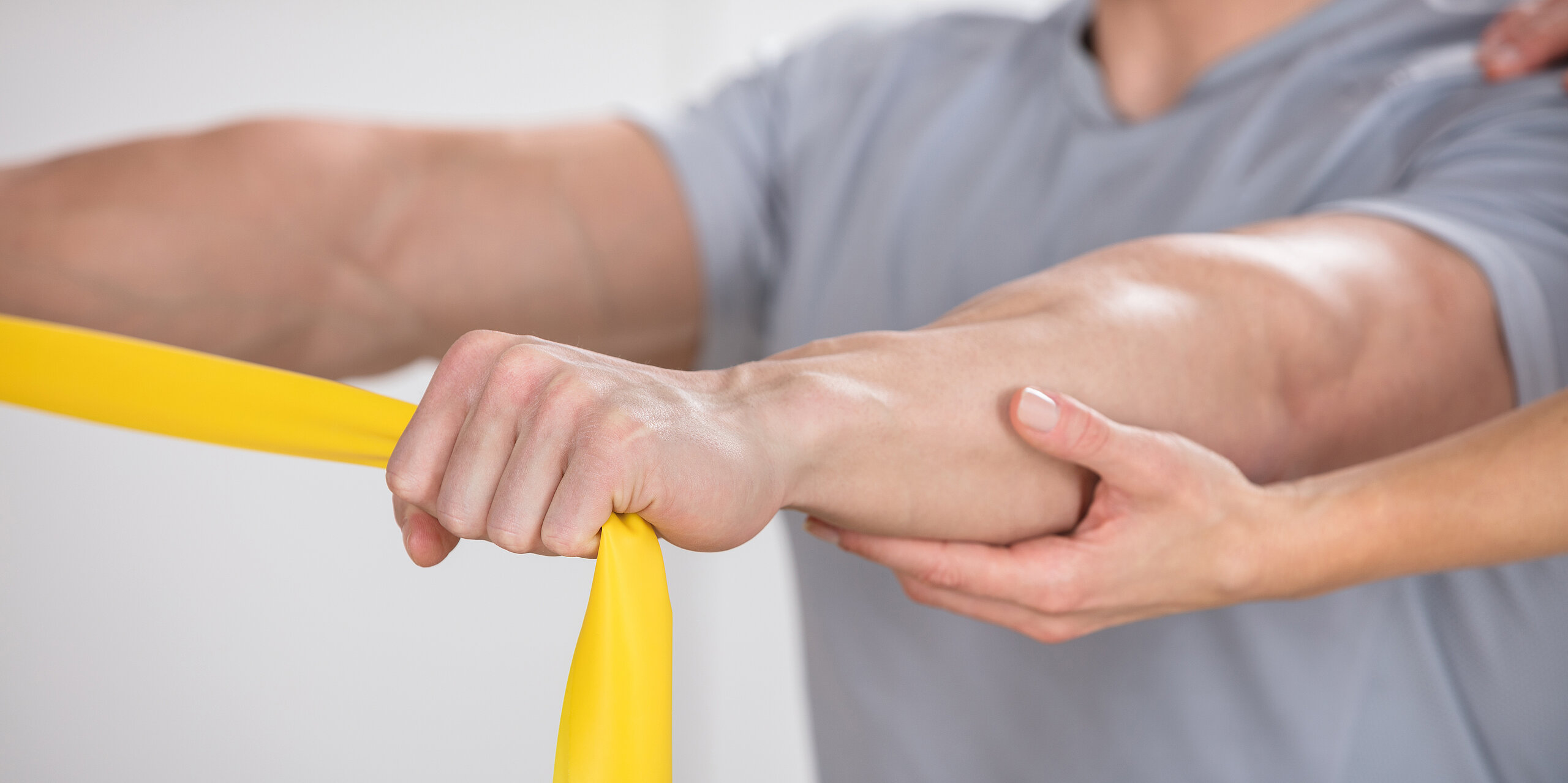 Ein Mann macht Übungen mit gelbem Theraband und wird dabei von einem Therapeuten unterstützt.