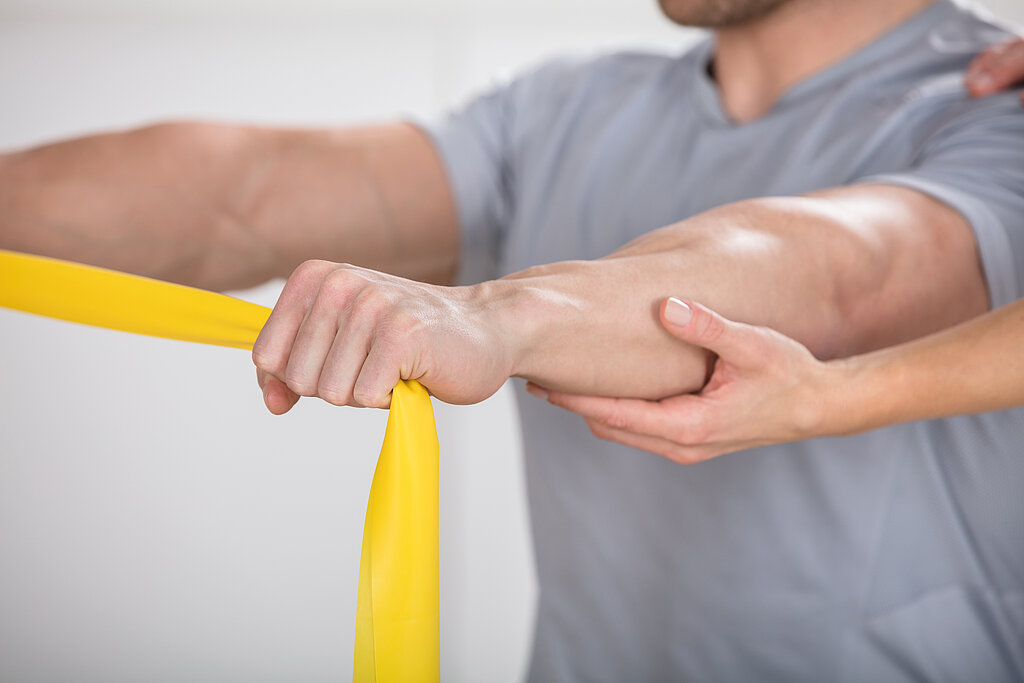 Physiotherapie mit Theraband Ein Mann macht Übungen mit gelbem Theraband und wird dabei von einem Therapeuten unterstützt.