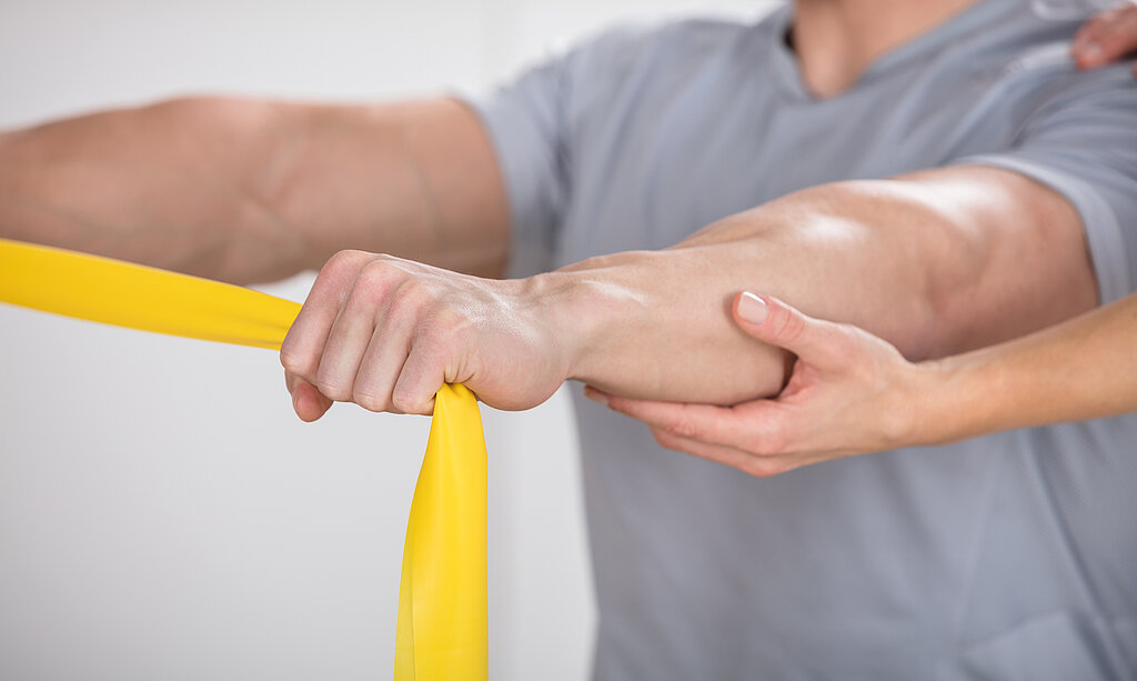 Ein Mann macht Übungen mit gelbem Theraband und wird dabei von einem Therapeuten unterstützt.