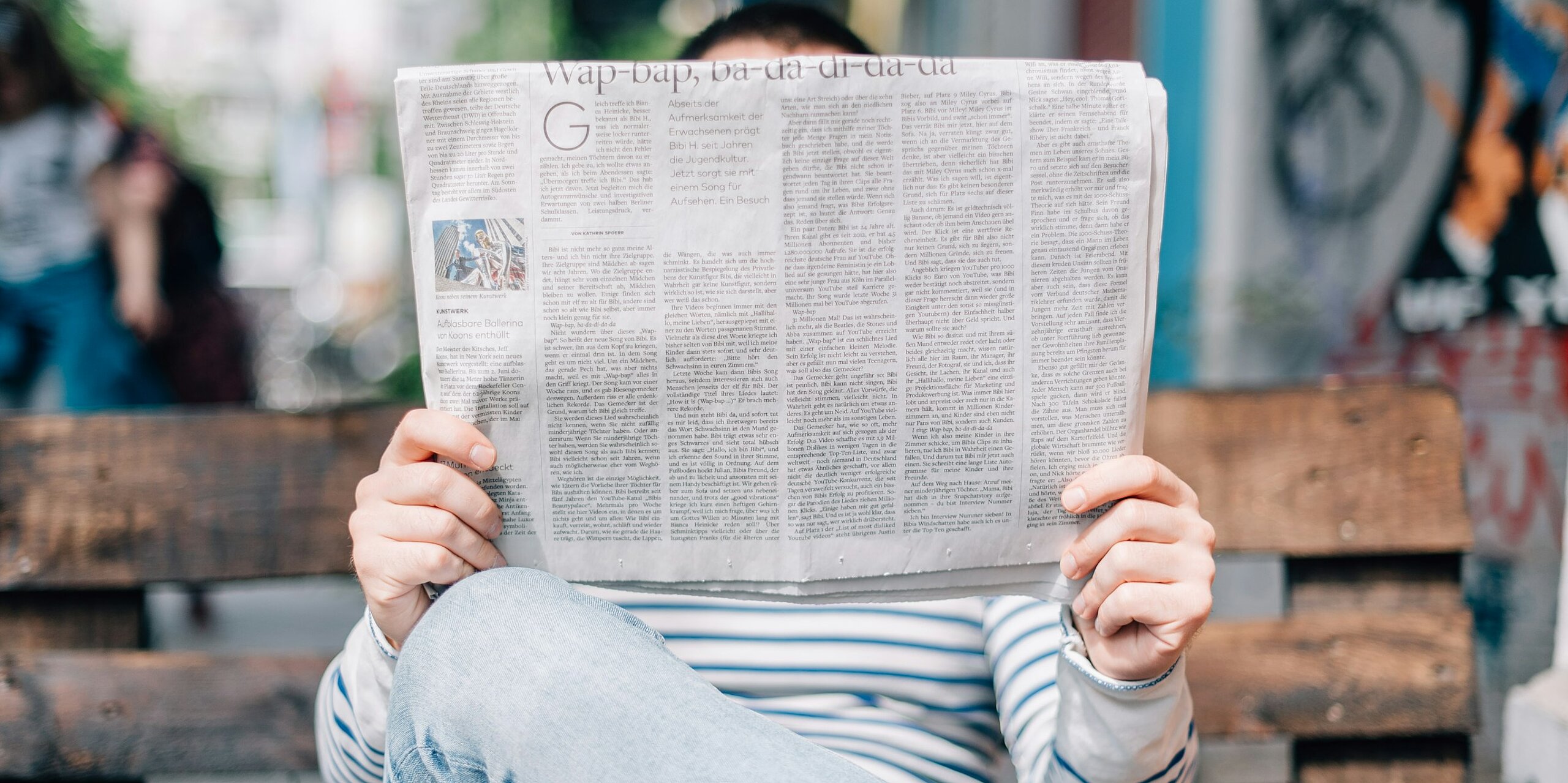 Ein Mensch ließt eine Zeitung auf einer Parkbank. Das Gesicht ist bedeckt von der Zeitung.