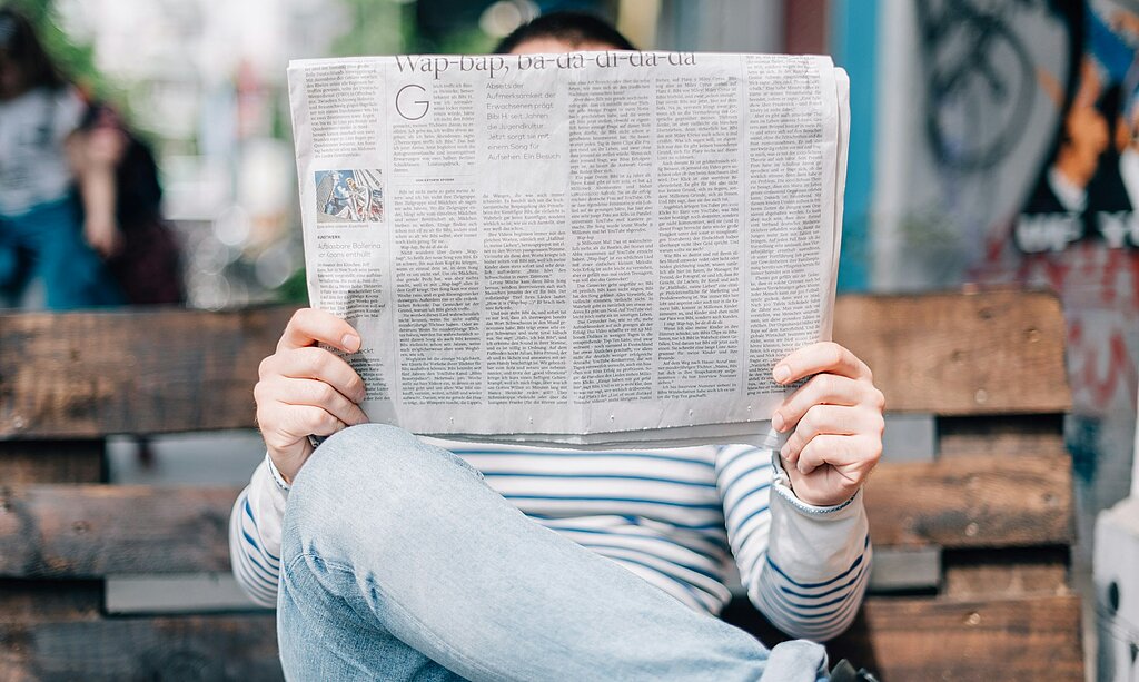 Ein Mensch ließt eine Zeitung auf einer Parkbank. Das Gesicht ist bedeckt von der Zeitung.