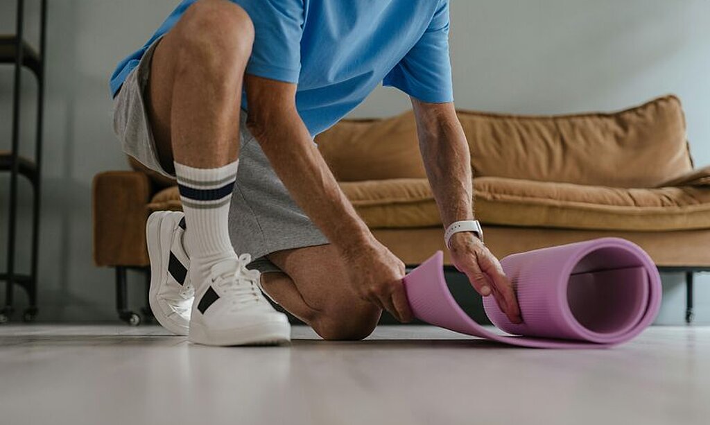 Ein Mann rollt eine Gymnastikmatte zuhause auf dem Boden aus.