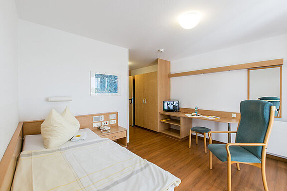 Das Bild zeigt ein eingerichtetes Zimmer mit Bett, Sessel, Spiegel, Kommoden und einem Fernseher.