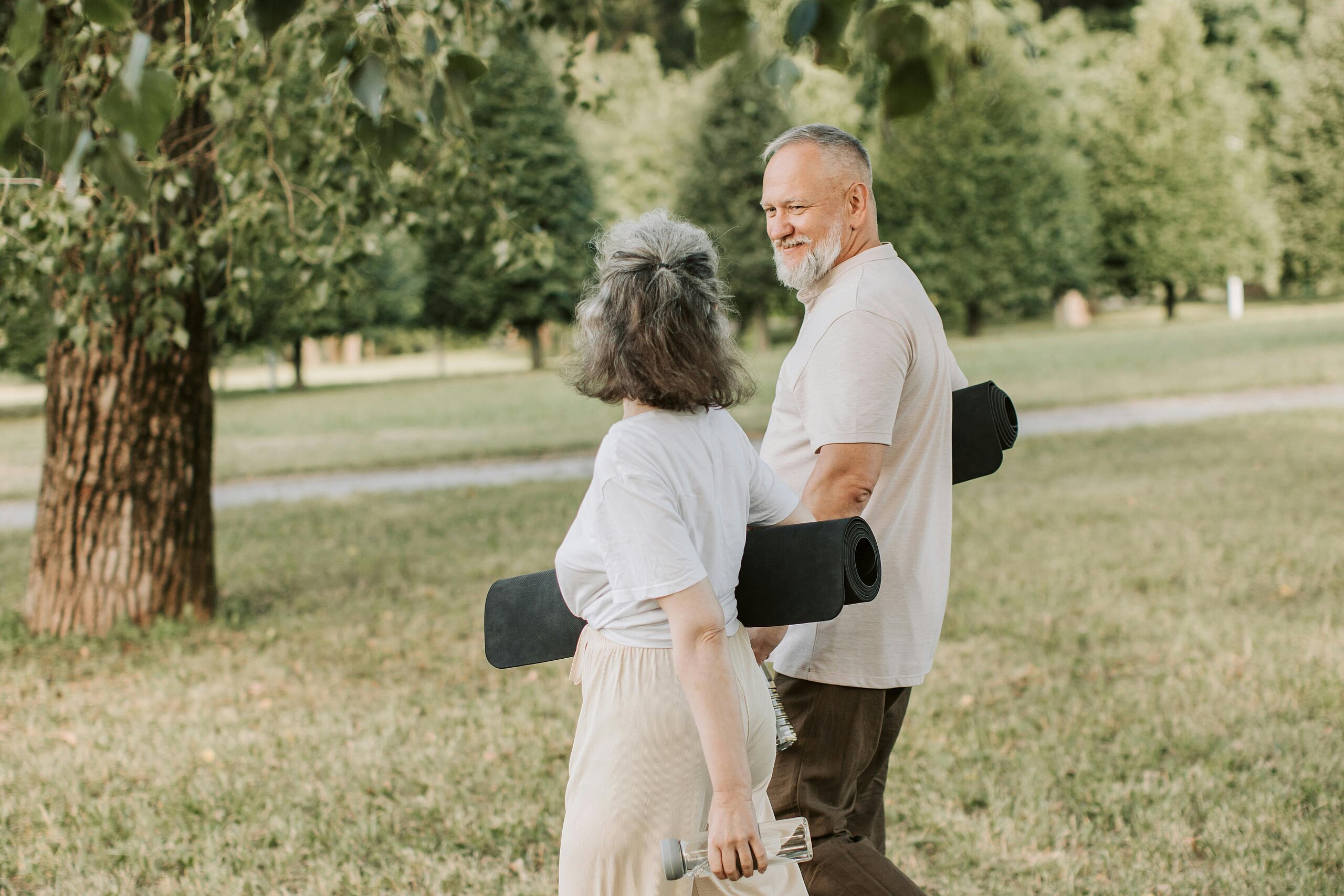 Eine ältere Frau und Mann gehen durch einen Park und gucken sich gegenseitig an. Unter ihrem Arm halten sie eine schwarze Yoga