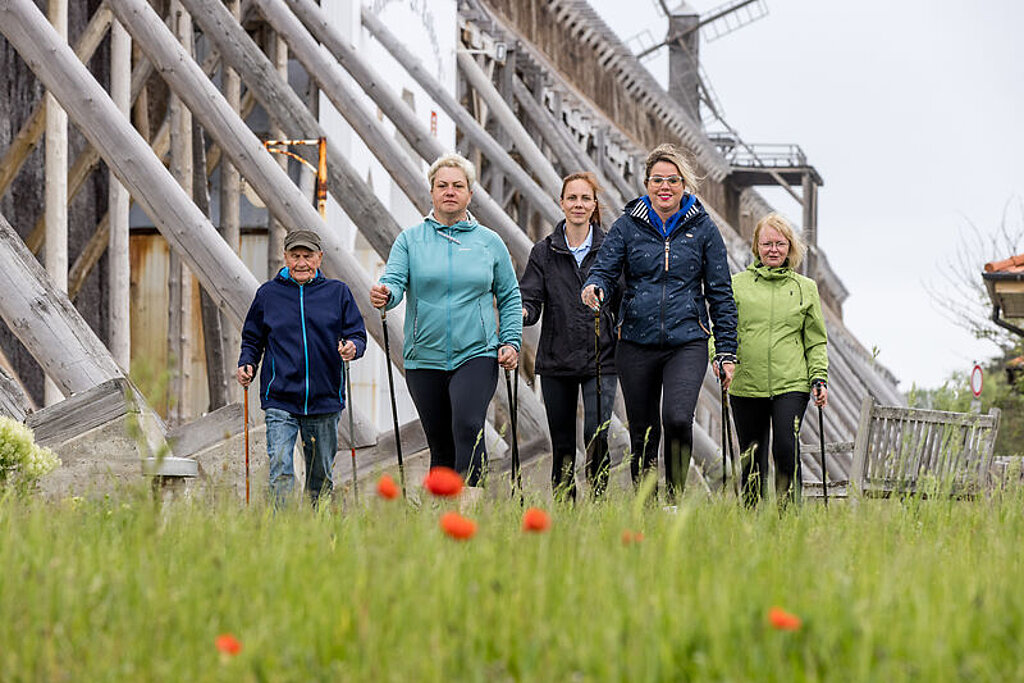 Fünf Personen laufen mit Walking-Stöcken in den Händen auf die Kamera zu, im Hintergrund befindet sich ein Gradierwerk.