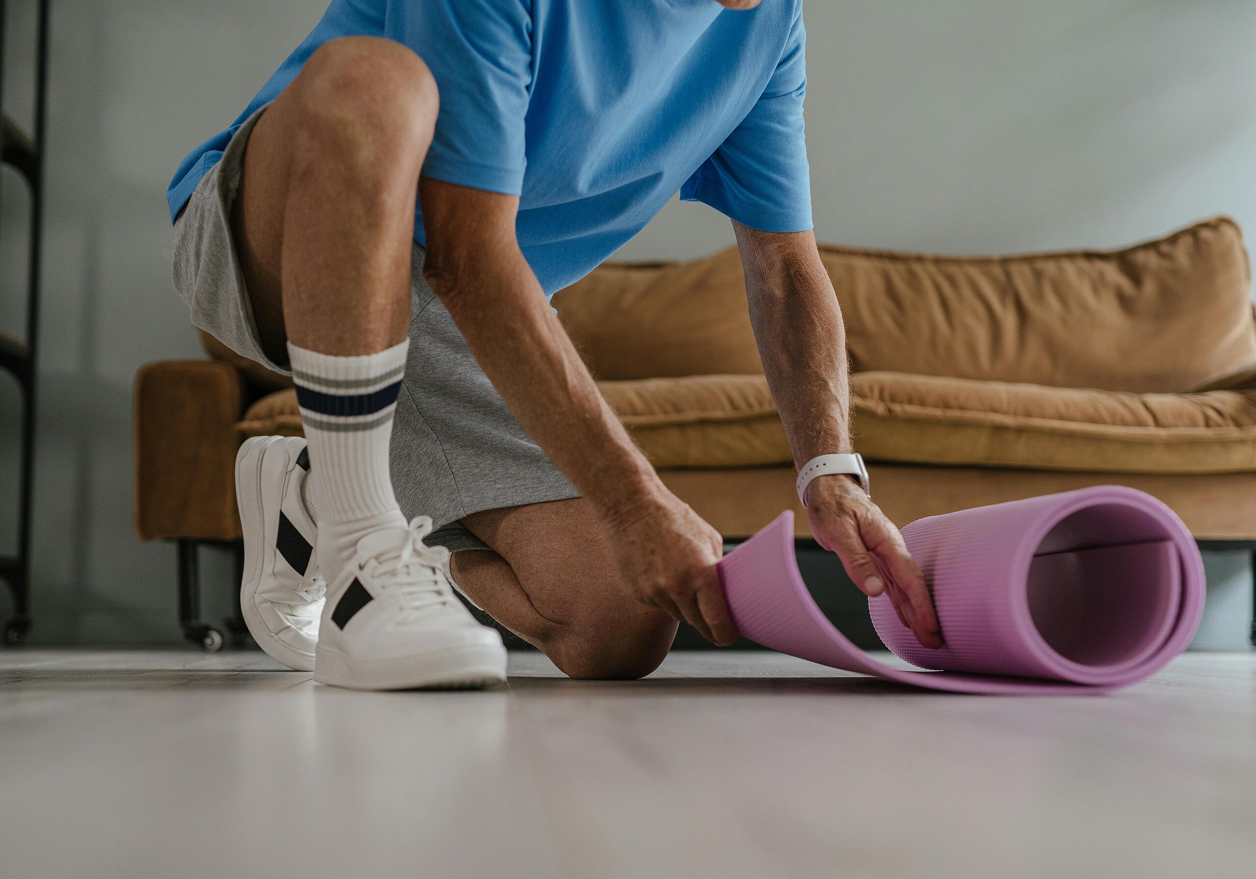 Ein älterer Mann in Sportkleidung rollt eine lila Yogamatte in einem Wohnzimmer aus, bereit für eine Trainingseinheit.