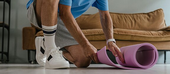 Ein älterer Mann in Sportkleidung rollt eine lila Yogamatte in einem Wohnzimmer aus, bereit für eine Trainingseinheit.