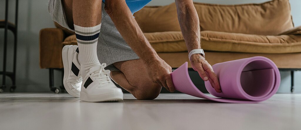 Ein älterer Mann in Sportkleidung rollt eine lila Yogamatte in einem Wohnzimmer aus, bereit für eine Trainingseinheit.
