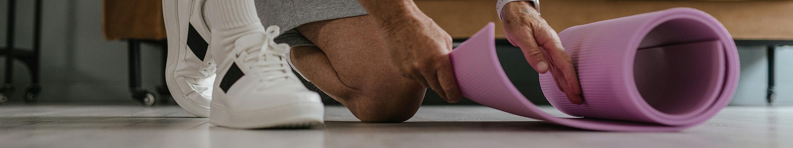 Ein älterer Mann in Sportkleidung rollt eine lila Yogamatte in einem Wohnzimmer aus, bereit für eine Trainingseinheit.