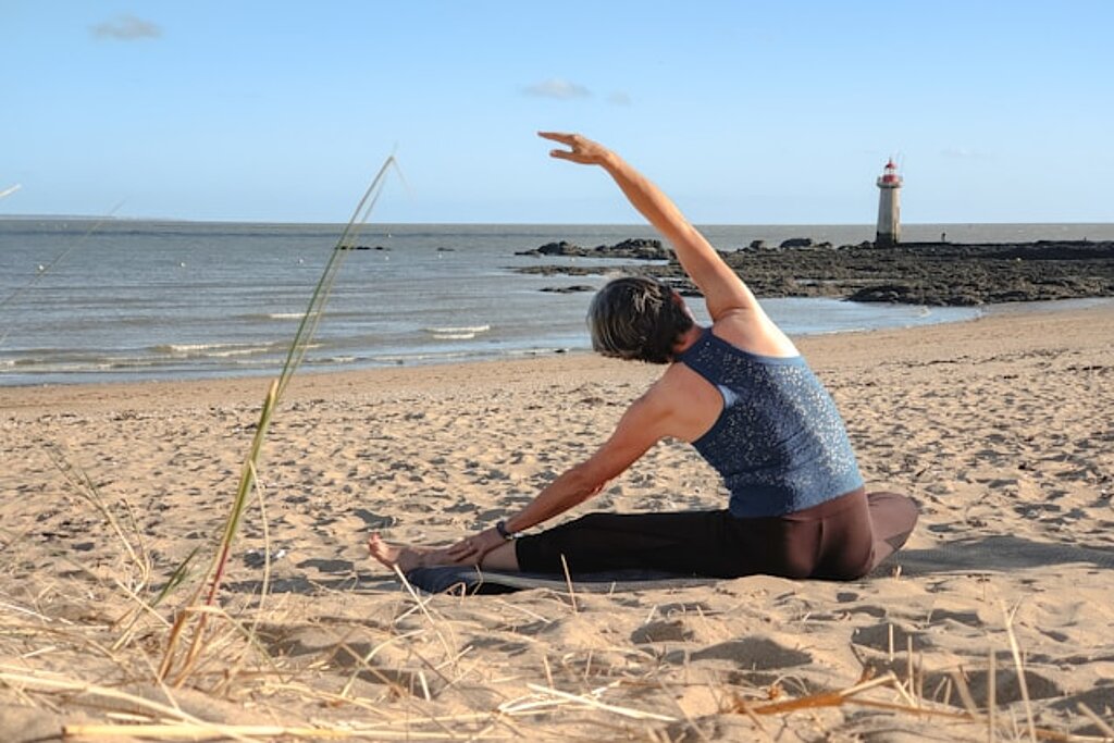 Eine Frau sitzt am Strand im Sand und macht Dehnübungen, im Hintergrund sieht man das Meer und einen Leuchtturm.