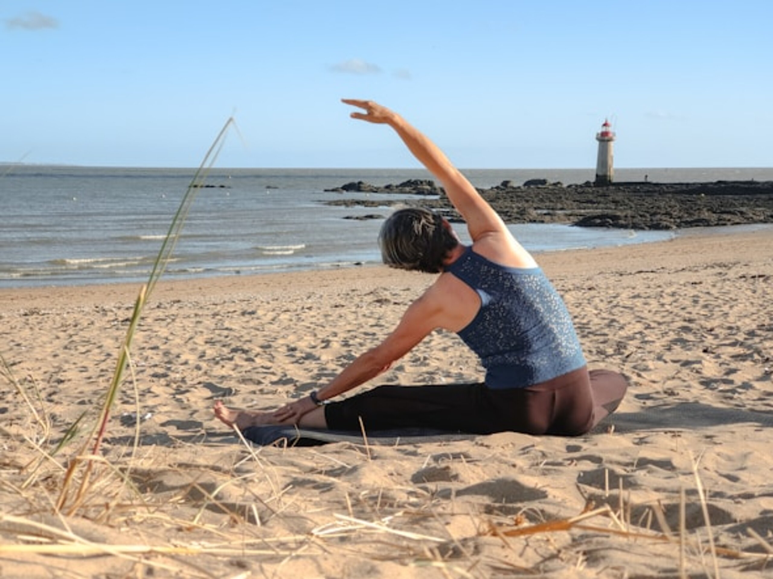 Eine Frau sitzt am Strand im Sand und macht Dehnübungen, im Hintergrund sieht man das Meer und einen Leuchtturm.