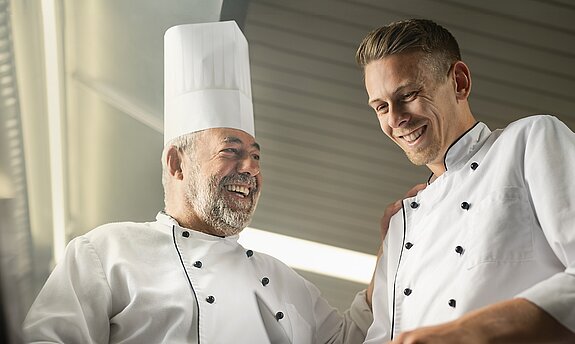 Ein alter Koch bringt einem jungen Koch etwas bei.