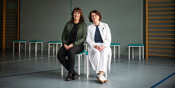 Dr. Ivonne Rudolph (links) und Dr. Bettine Bilsing sitzen in einer Sporthalle auf Stühlen.