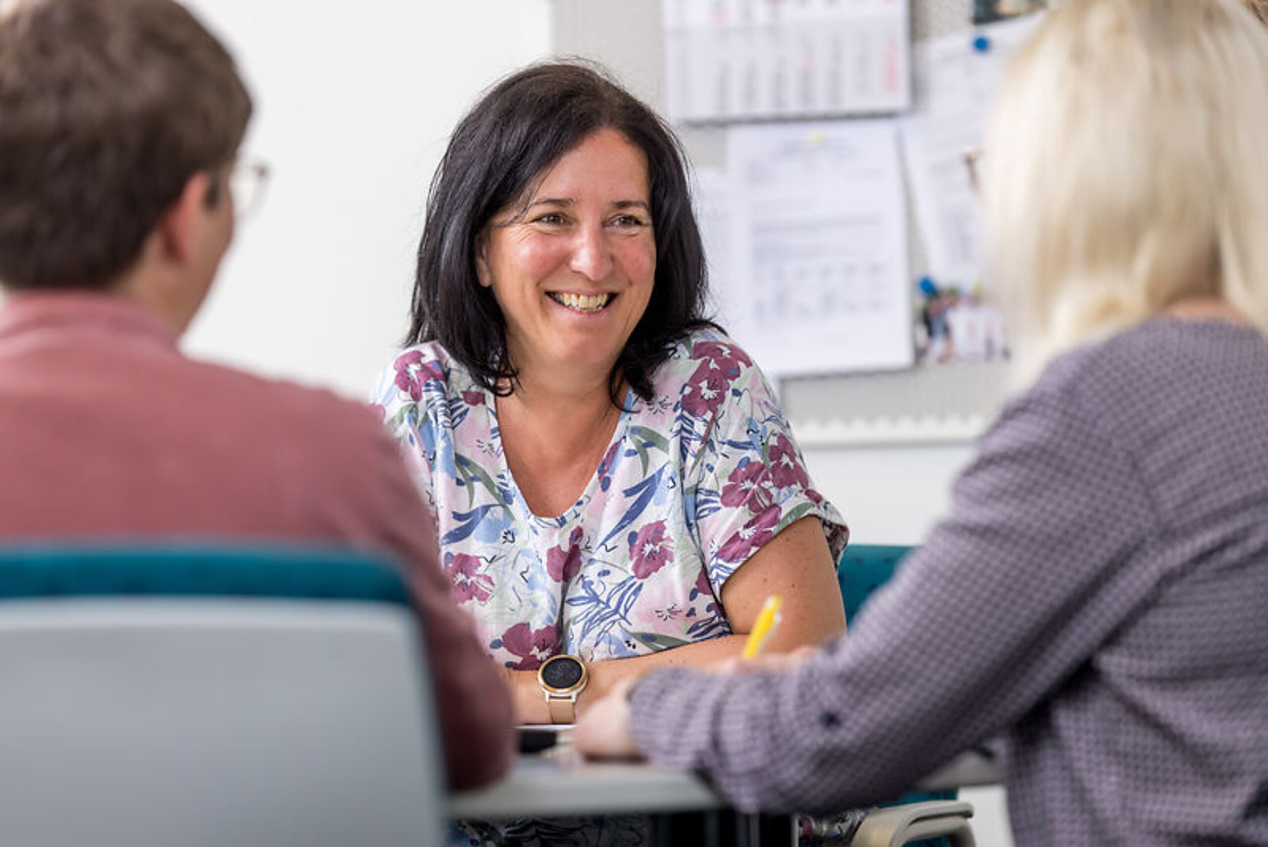 Gespräch Patienten Eine Mitarbeiterin unterhält sich freundlich mit zwei Patienten am Schreibtisch.