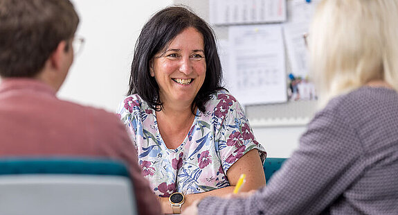 Gespräch Patienten Eine Mitarbeiterin unterhält sich freundlich mit zwei Patienten am Schreibtisch.