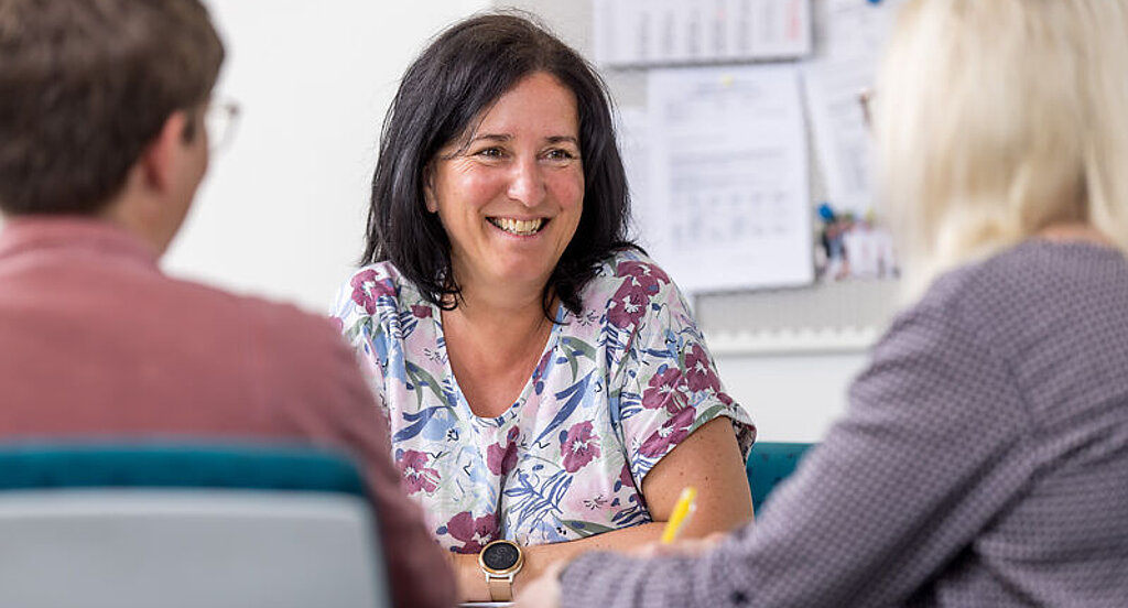 Gespräch Patienten Eine Mitarbeiterin unterhält sich freundlich mit zwei Patienten am Schreibtisch.