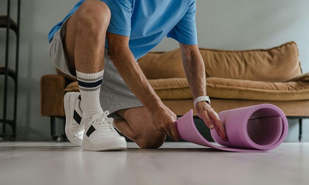 Ein Mann rollt zuhause eine Trainingsmatte auf dem Boden aus.