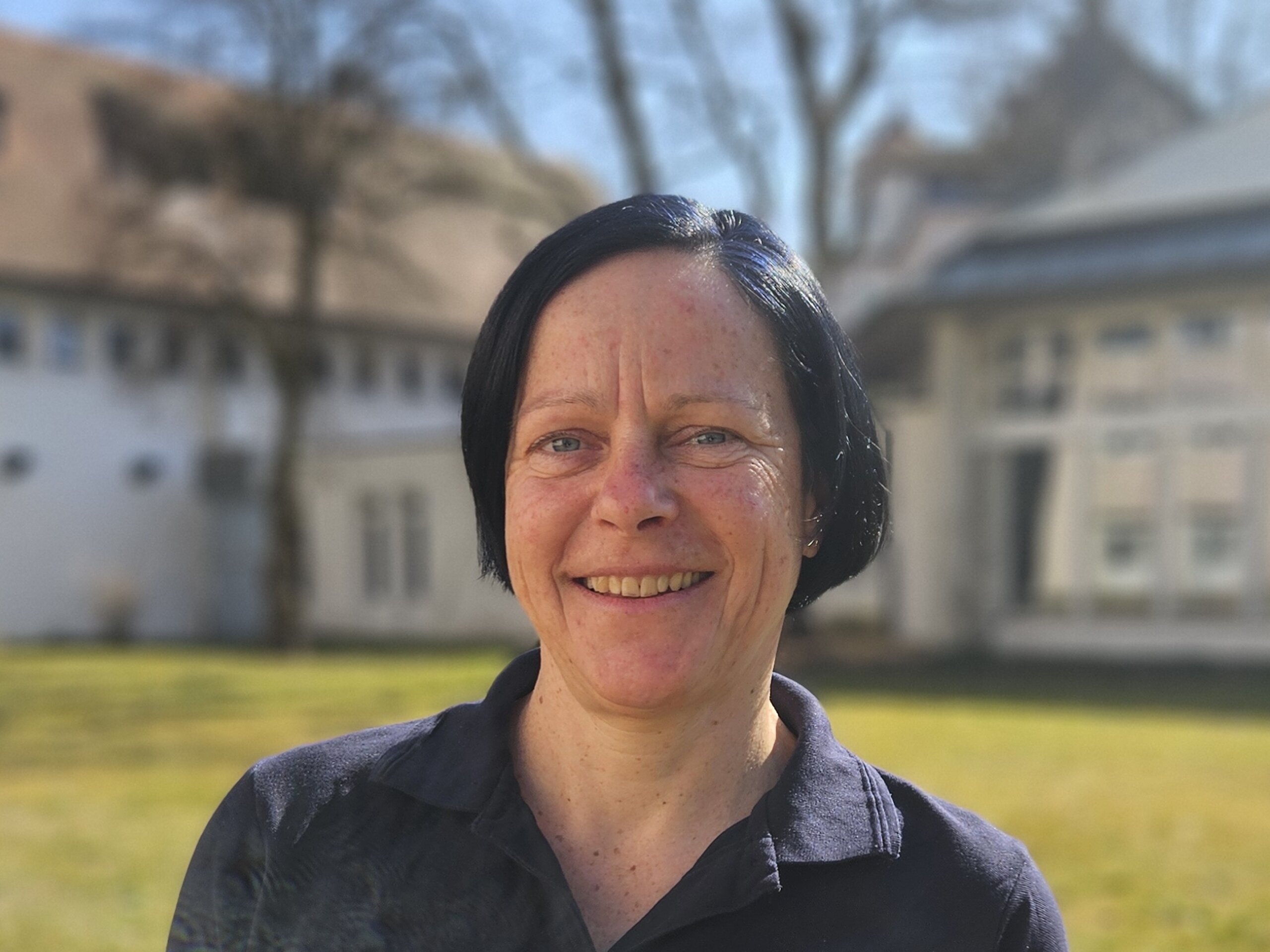 Eine Therapeutin der KIH im Freien auf einer Wiese mit einem Gebäude im Hintergrund und trägt dunkle Therapie-Arbeitskleidung.